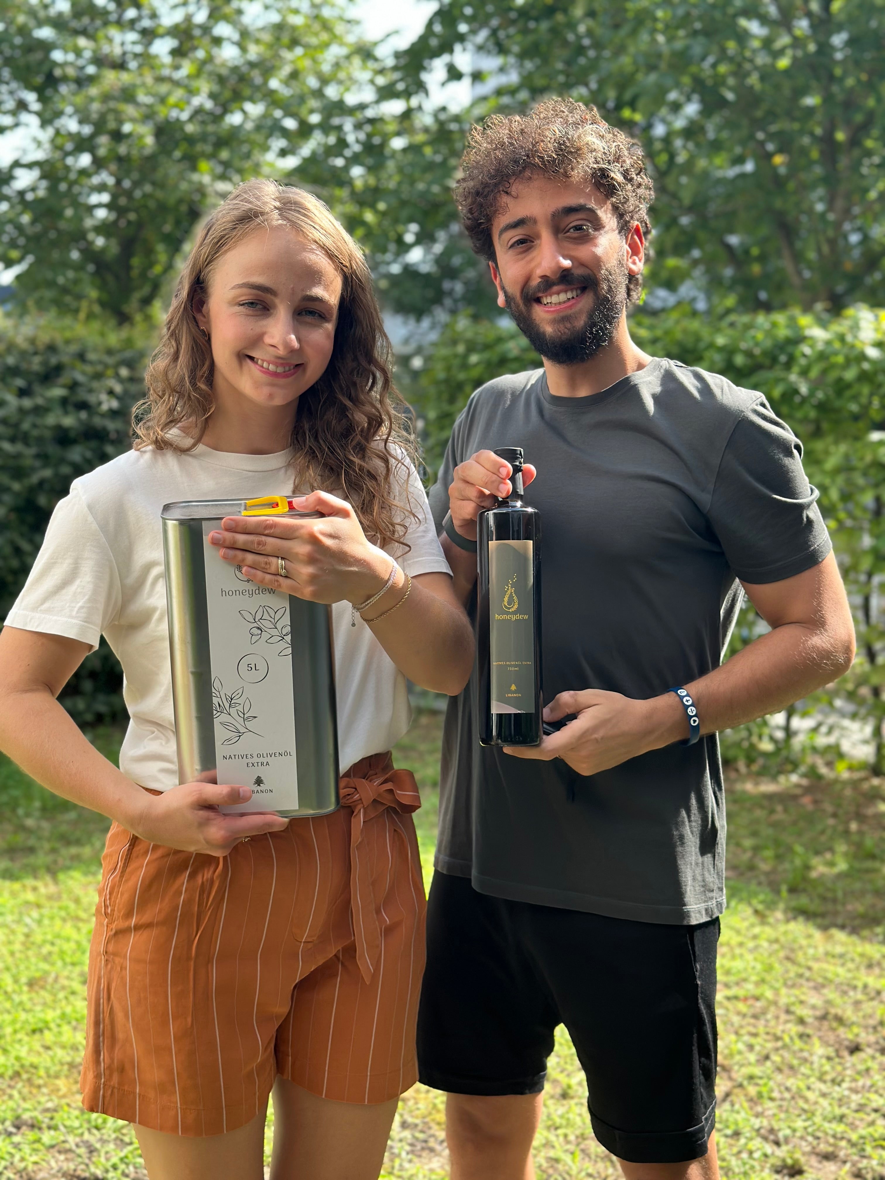 Eine lächelnde Frau hält einen 5-Liter-Kanister mit extra nativem Olivenöl, während ein lächelnder Mann eine 750-ml-Glasflasche Olivenöl präsentiert, beide stehen im Grünen