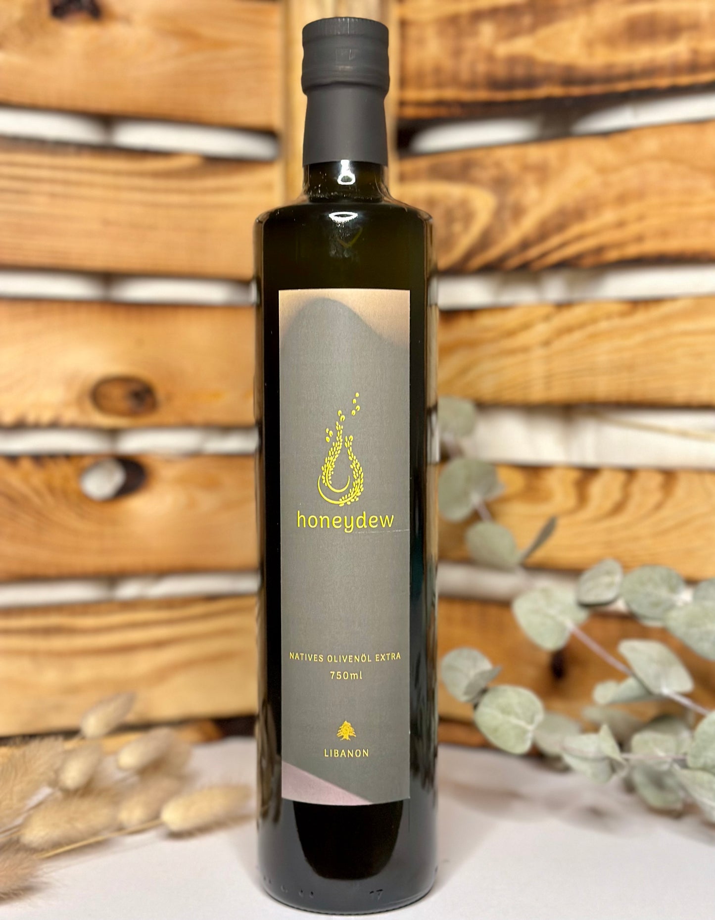 Eine 750-ml-Flasche libanesisches natives Olivenöl extra von "honeydew" steht vor einem rustikalen Holzhintergrund, umgeben von Trockenblumen und Eukalyptusblättern.