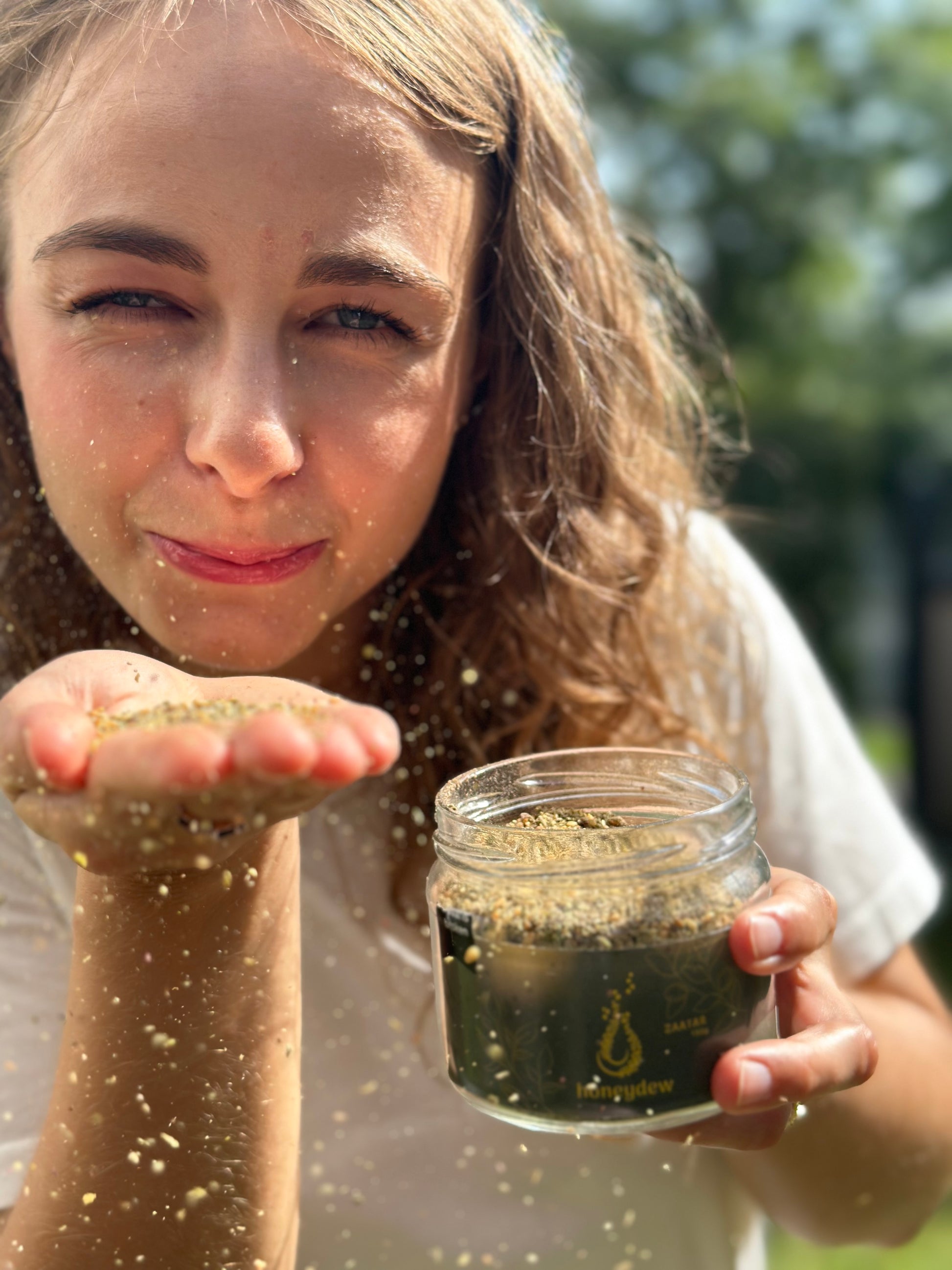 Junge Frau hält ein Glas libanesische Zaatar-Mischung und lässt das Gewürz aus ihrer Hand rieseln, vor sonniger, grüner Kulisse