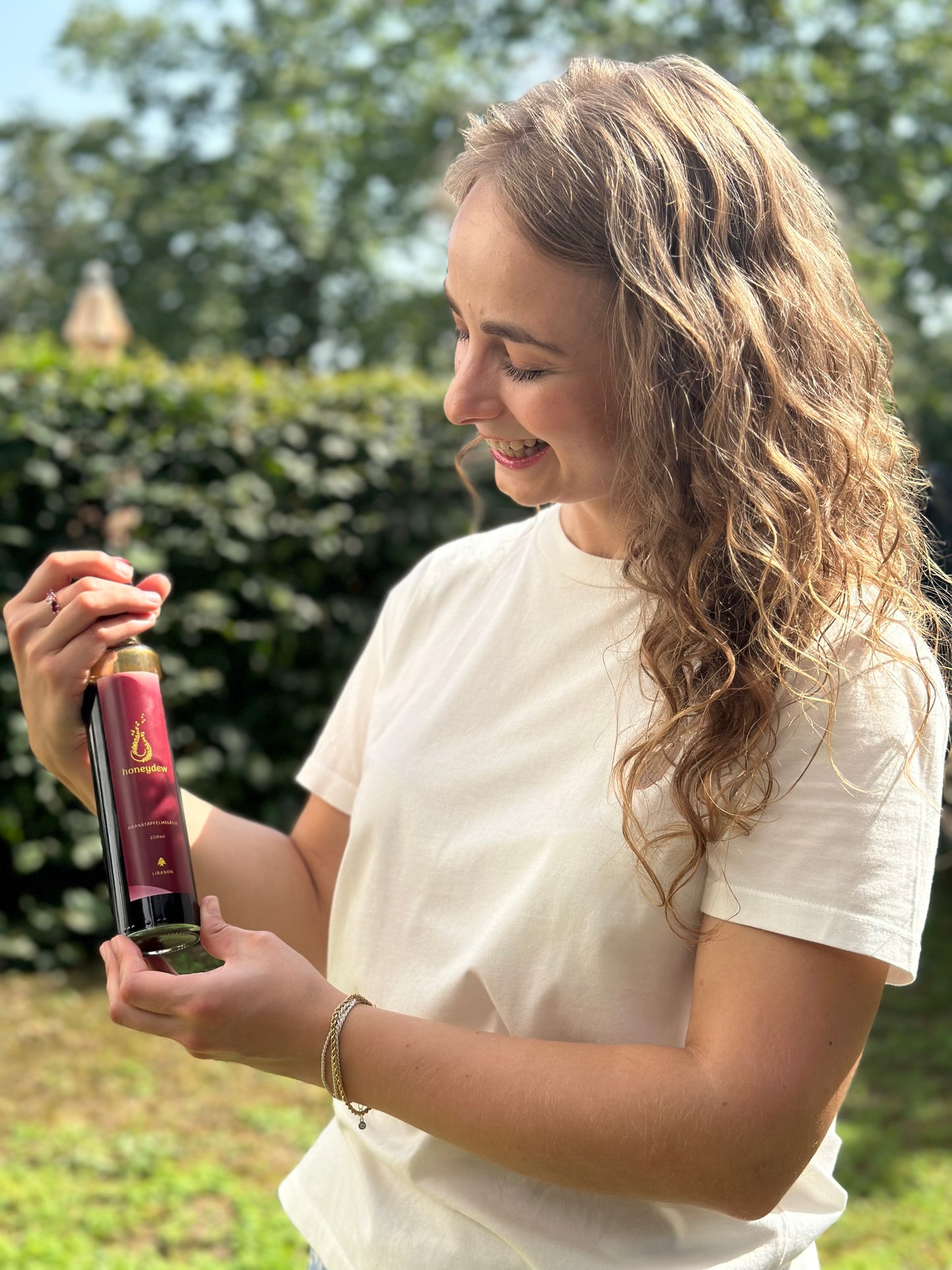 Junge Frau hält eine Flasche honeydew Granatapfelmelasse mit rotem Etikett und goldenem Logo vor grünem, sonnigem Hintergrund