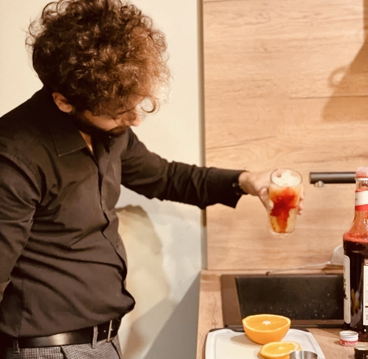 Ein Mann in schwarzem Hemd bereitet ein Getränk mit Eis und Granatapfelmelasse zu, daneben eine aufgeschnittene Orange auf einem Tablett.