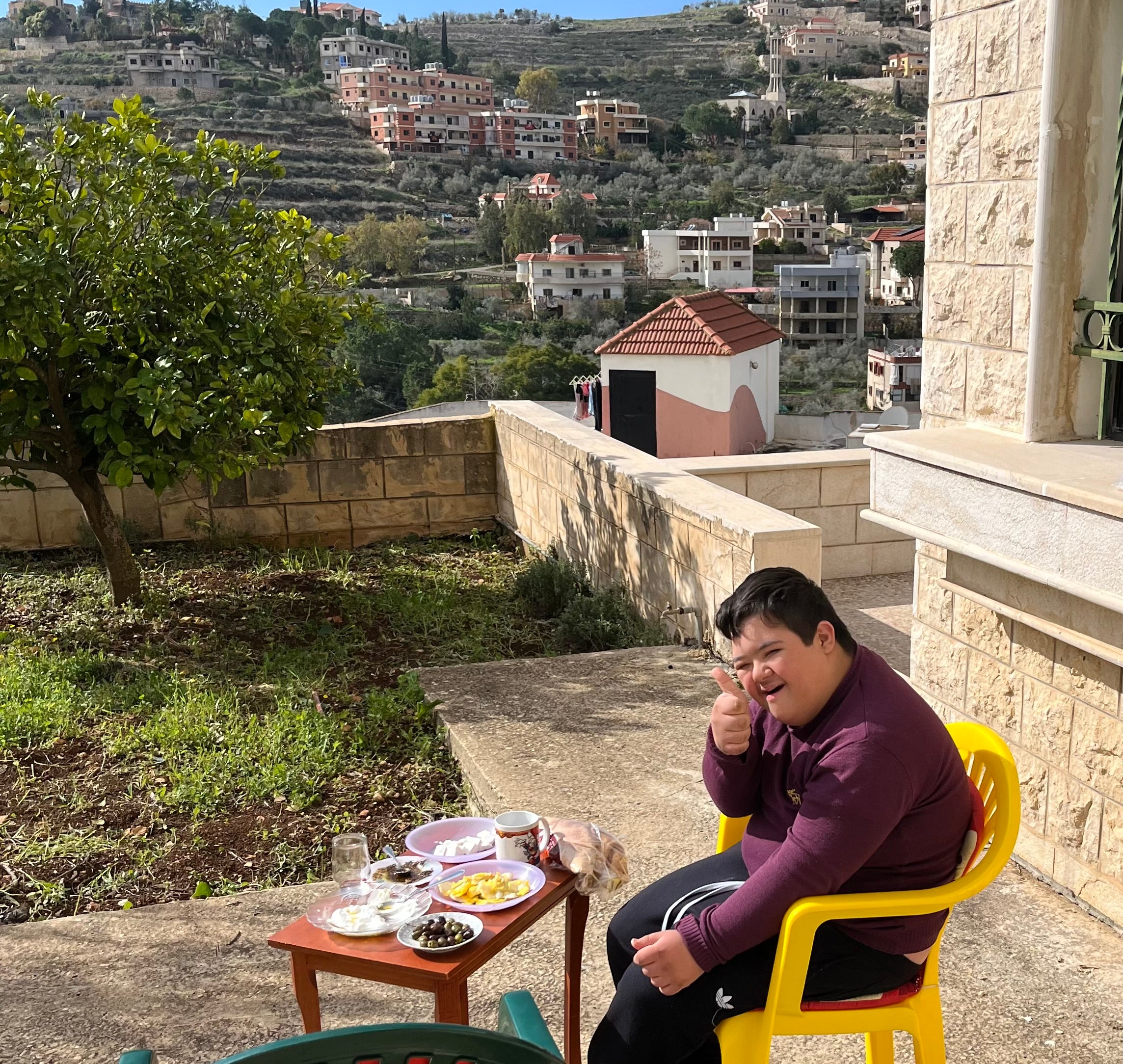 Junger Mann mit Down-Syndrom sitzt auf einem gelben Stuhl und zeigt fröhlich einen Daumen hoch neben einem Tisch mit traditionellem libanesischen Frühstück, vor einer Landschaft mit Hügeln und Häusern