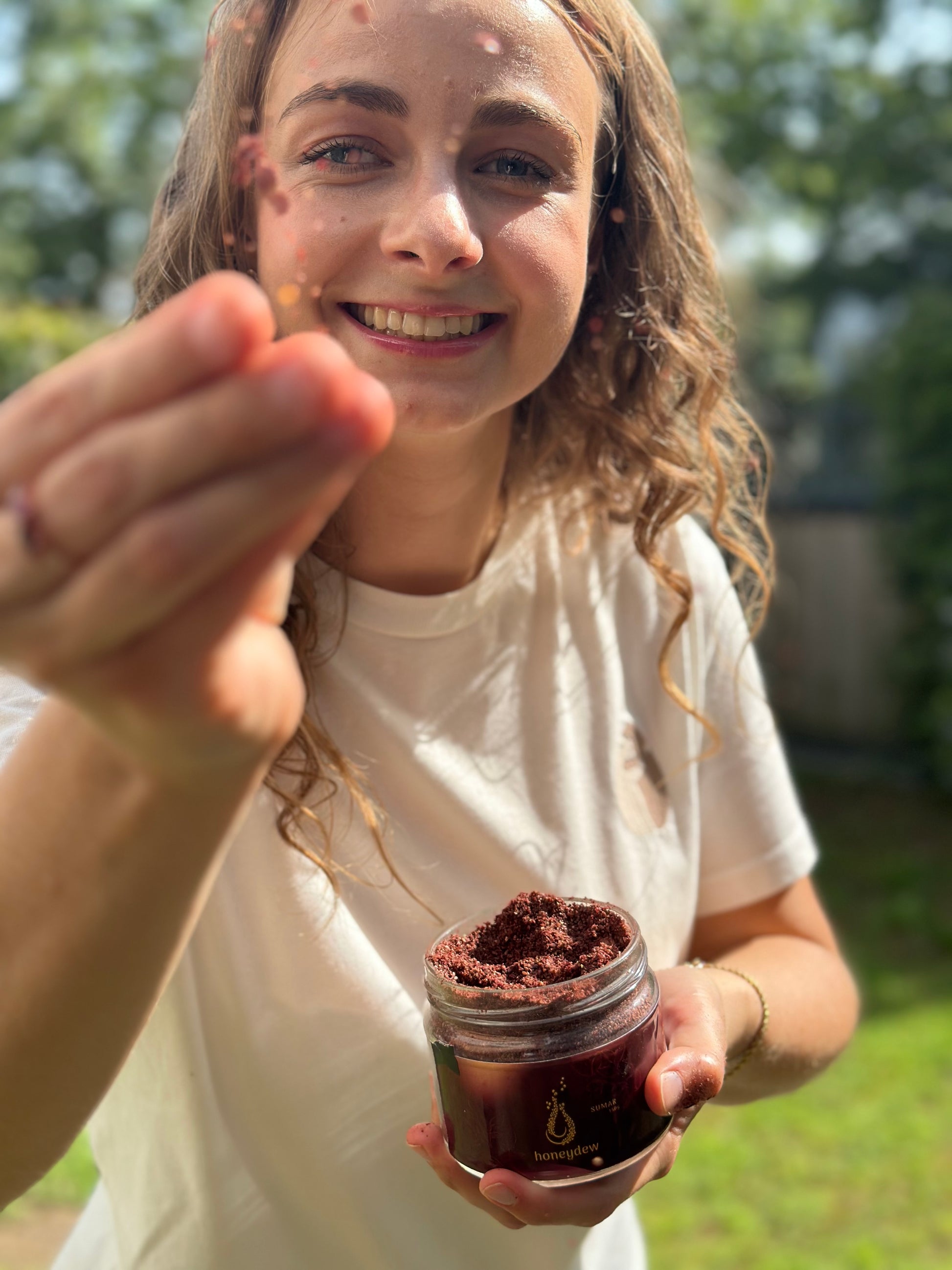 Eine junge Frau mit langen, welligen Haaren und einem strahlenden Lächeln hält ein Glas Honeydew libanesischen Sumach in der Hand. Mit der anderen Hand streut sie das tiefrote Gewürz spielerisch in die Kamera
