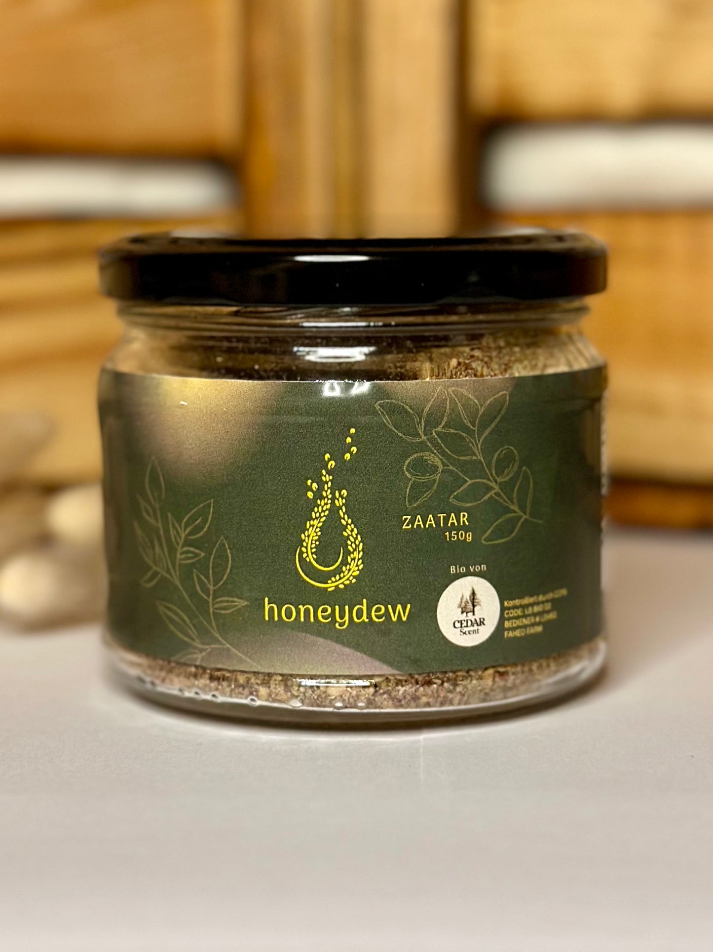 Ein stilvolles Glas "Honeydew Zaatar" mit schwarzem Deckel steht im Fokus des Bildes. Das edle, dunkelgrüne Etikett mit goldfarbenem Logo und filigranen Pflanzenillustrationen vermittelt Qualität und Natürlichkeit. Die klare Beschriftung zeigt „ZAATAR 150g“ und das Bio-Siegel von Cedar Scent unterstreicht den authentischen Ursprung des Produkts. Der warme Hintergrund mit Holzdetails rundet die Szene harmonisch ab und vermittelt ein Gefühl von Handwerkskunst und Tradition.