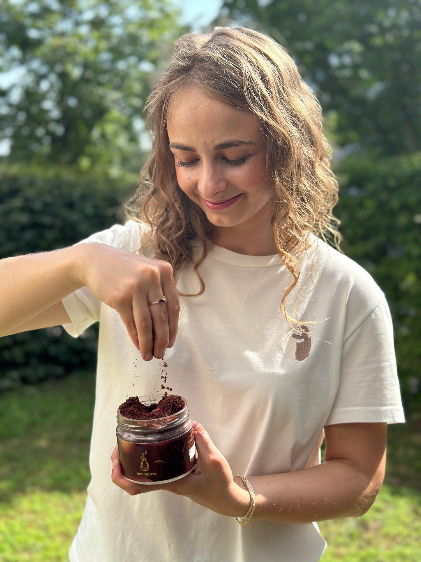 Eine junge Frau mit welligen blonden Haaren und einem warmen Lächeln hält ein Glas Honeydew libanesischer Sumach in der Hand und lässt das aromatische Gewürz sanft zwischen ihren Fingern rieseln.
