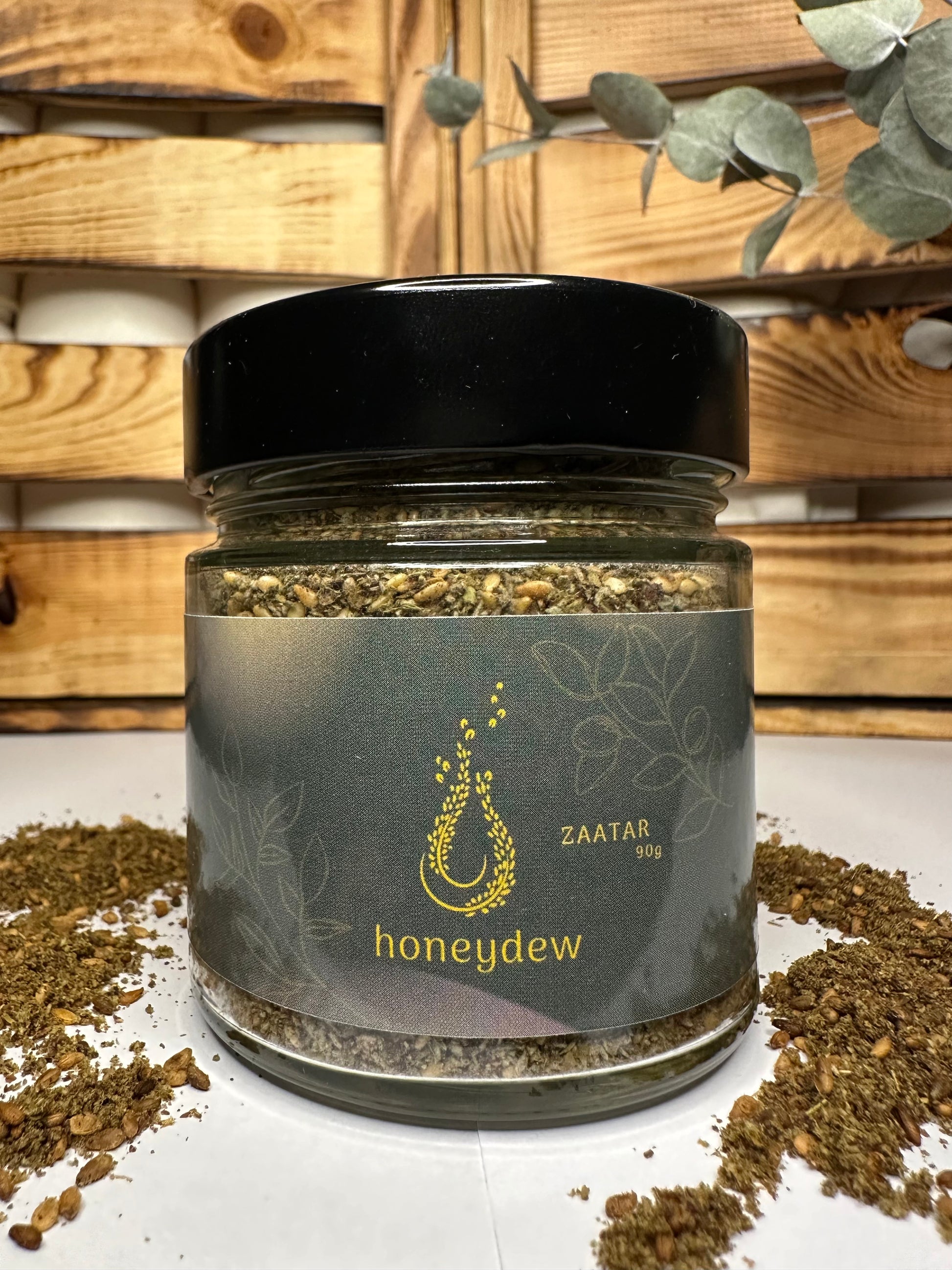 Libanesische Honeydew Zaatar Mischung im 90g-Glas mit schwarzem Deckel, goldfarbenem Logo und verstreutem Zaatar vor rustikalem Holzhintergrund