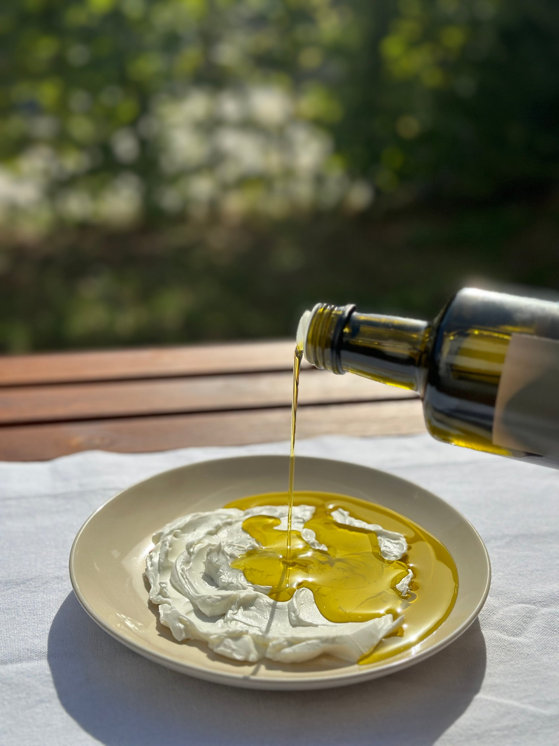 Eine Nahaufnahme zeigt, wie goldgelbes Olivenöl aus einer dunklen Glasflasche auf einen Teller mit cremigem, weißem Joghurt gegossen wird. 