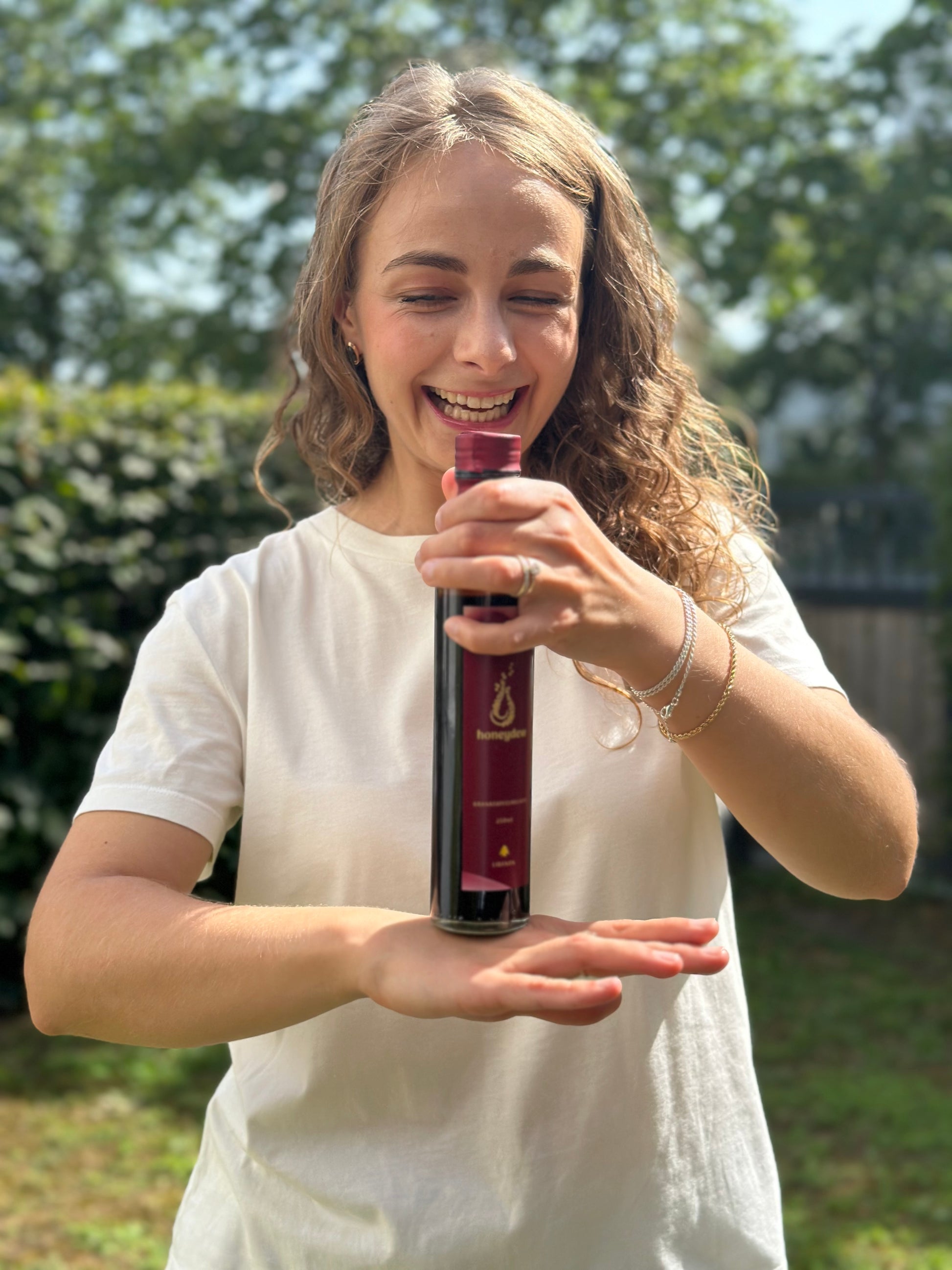 Junge Frau hält eine Flasche honeydew Granatapfelmelasse, rotes Etikett mit goldener Schrift, vor grünem, sonnigem Hintergrund