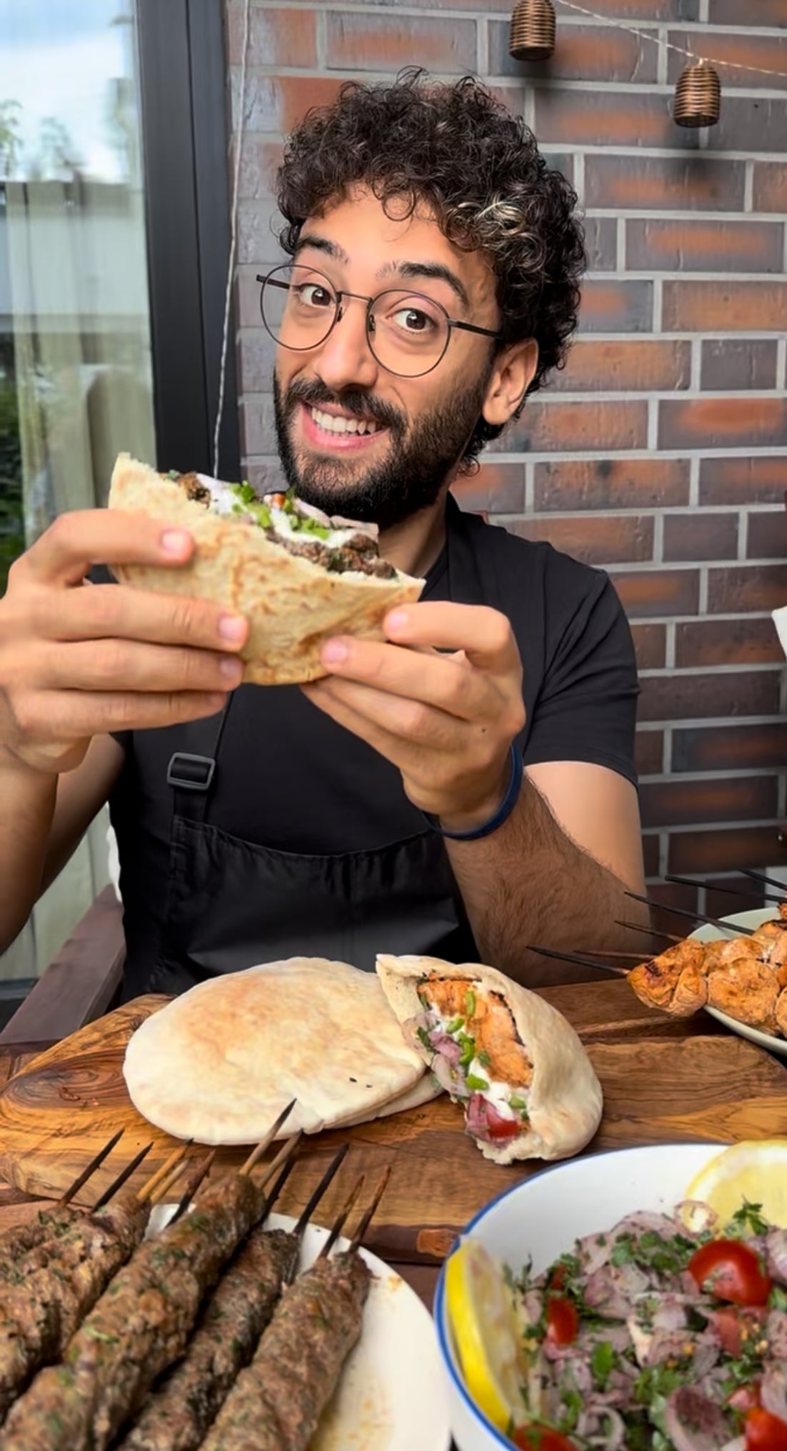 Person hält ein libanesisches Pita-Sandwich mit gegrilltem Fleisch und frischem Salat, umgeben von Spießen, Fladenbrot und Taboulé auf einem Holztisch.