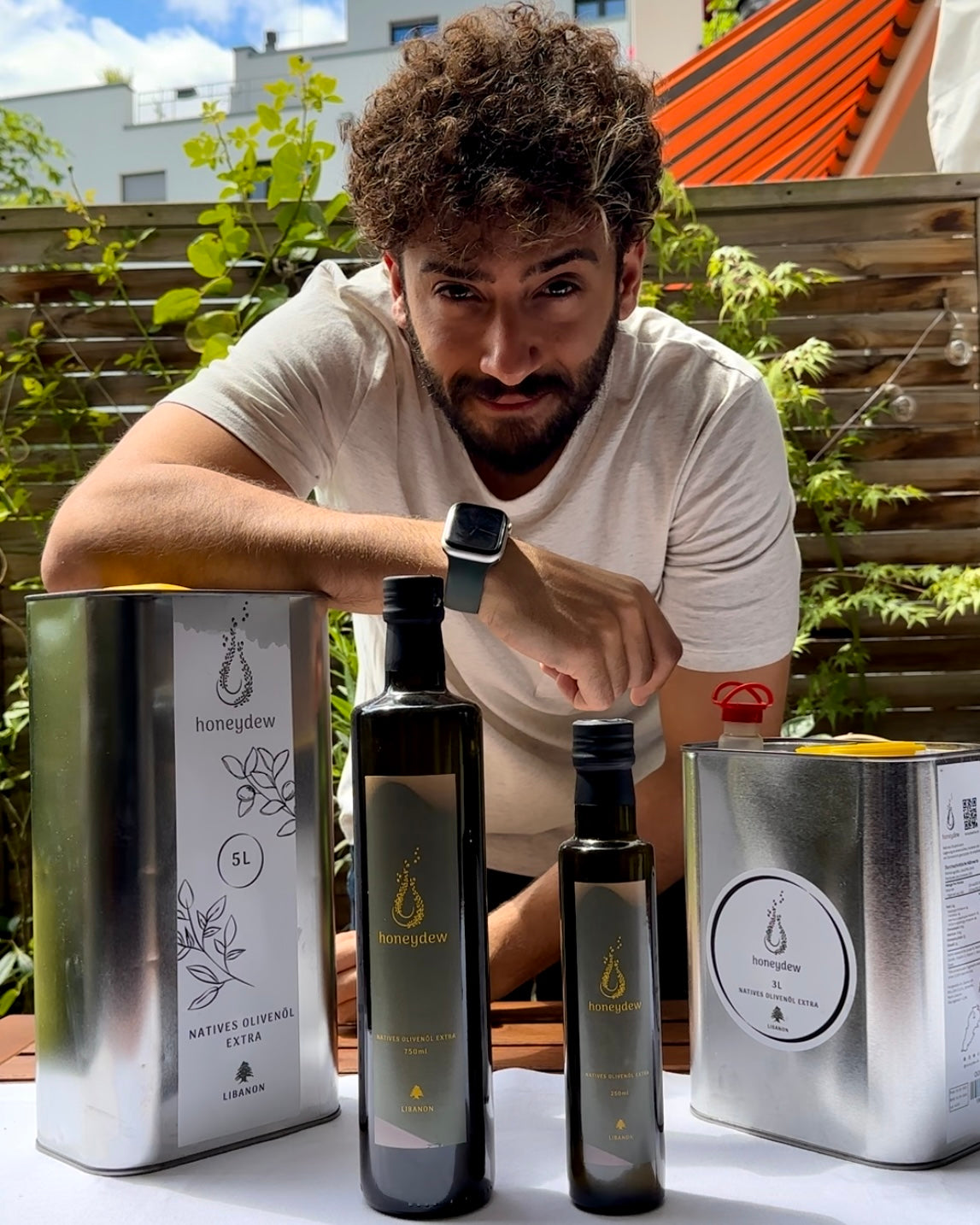 Gründer präsentiert Honeydew Natives Olivenöl Extra-Produkte, darunter 5L- und 3L-Kanister sowie 750ml- und 250ml-Flaschen, aus dem Libanon, in einer sonnigen Außenumgebung mit viel Grün