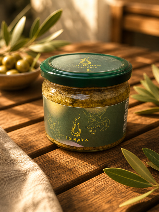 Honeydew Tapenade Grün 250g im Glasglas mit grünem Deckel auf rustikalem Holztablett, umgeben von frischen Olivenzweigen und einer Schale grüner Oliven im warmen Gegenlicht