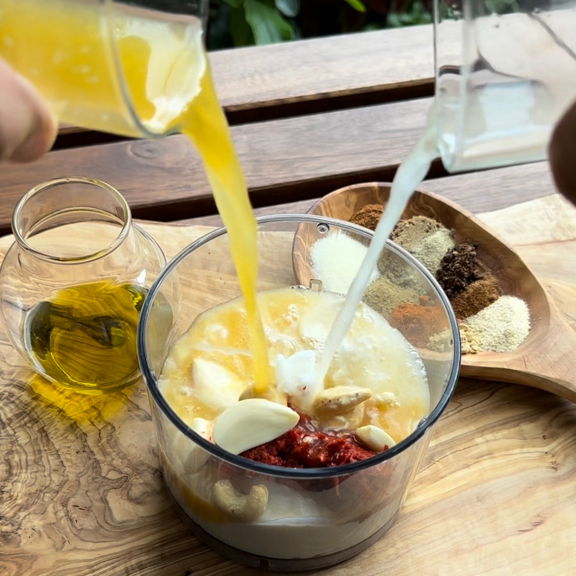 Nahaufnahme einer Küchenzubereitung mit frischem Zitronensaft und Kokosmilch, die in einen Mixer mit Cashewkernen, Knoblauch und Gewürzen gegossen werden, daneben ein Glas Olivenöl und eine Holzschale mit Gewürzen