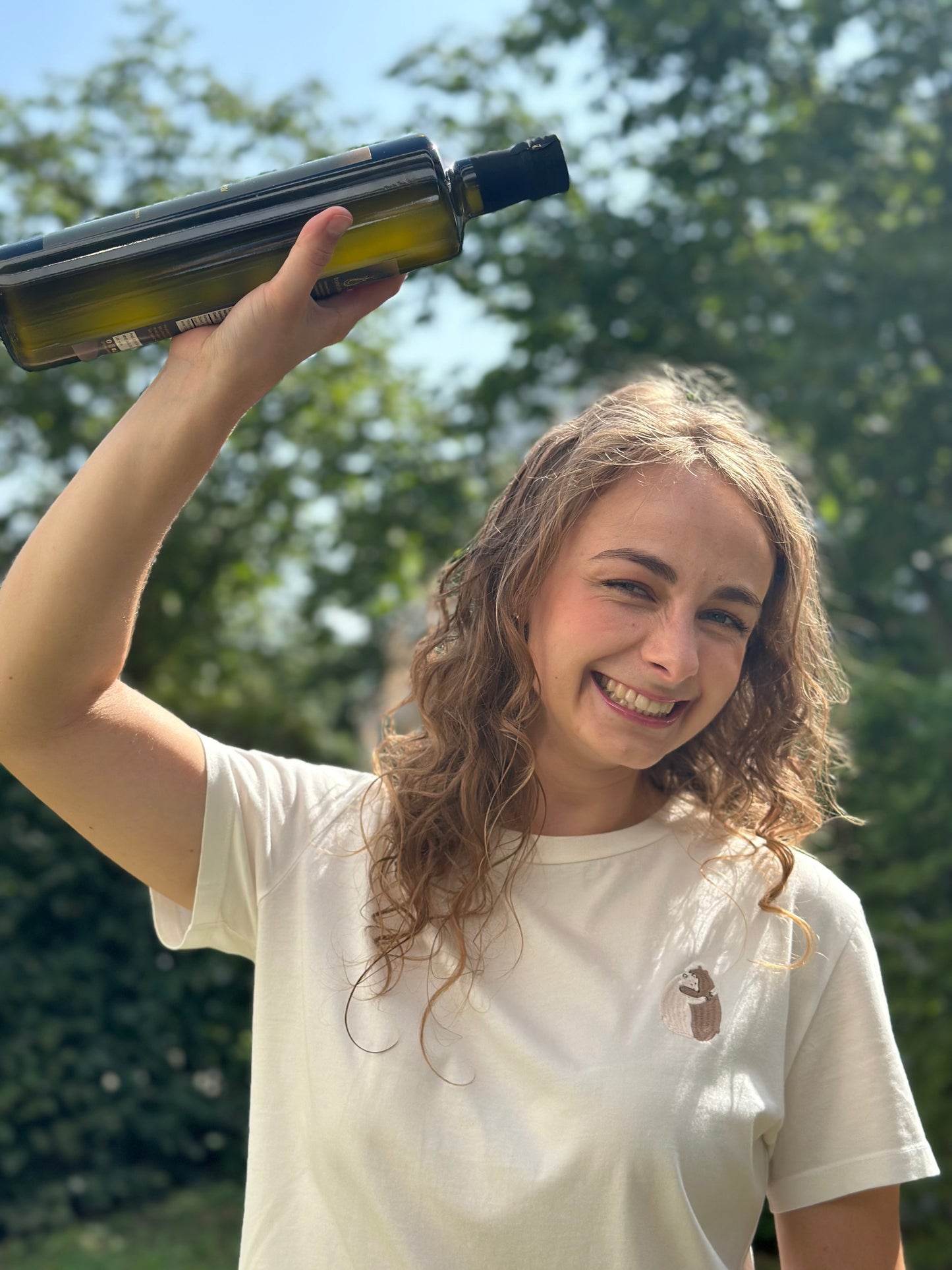 Junge Frau mit lockigen Haaren und weißem T-Shirt hält eine Flasche libanesisches natives Olivenöl extra über ihren Kopf und lächelt in die Kamera, vor einem sonnigen, grünen Hintergrund