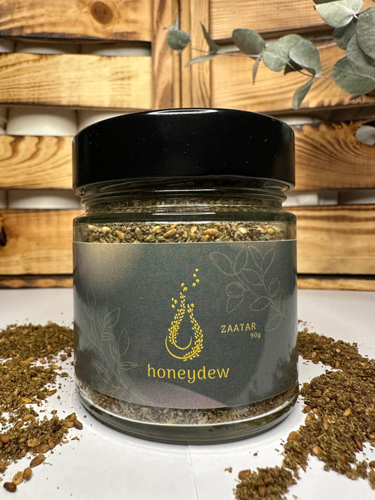 Libanesische Honeydew Zaatar Mischung im 90g-Glas mit schwarzem Deckel, goldfarbenem Logo und verstreutem Zaatar vor rustikalem Holzhintergrund