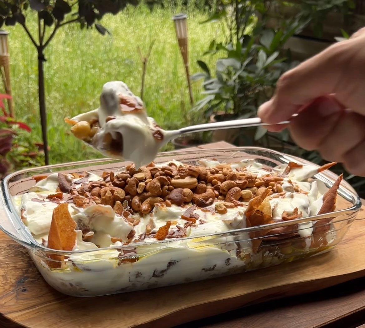 Libanesisches Fatteh-Gericht mit cremigem Joghurt, knusprigem Brot, gerösteten Nüssen und einem Schuss nativem Olivenöl extra, serviert in einer Glasform auf einem Holztisch.