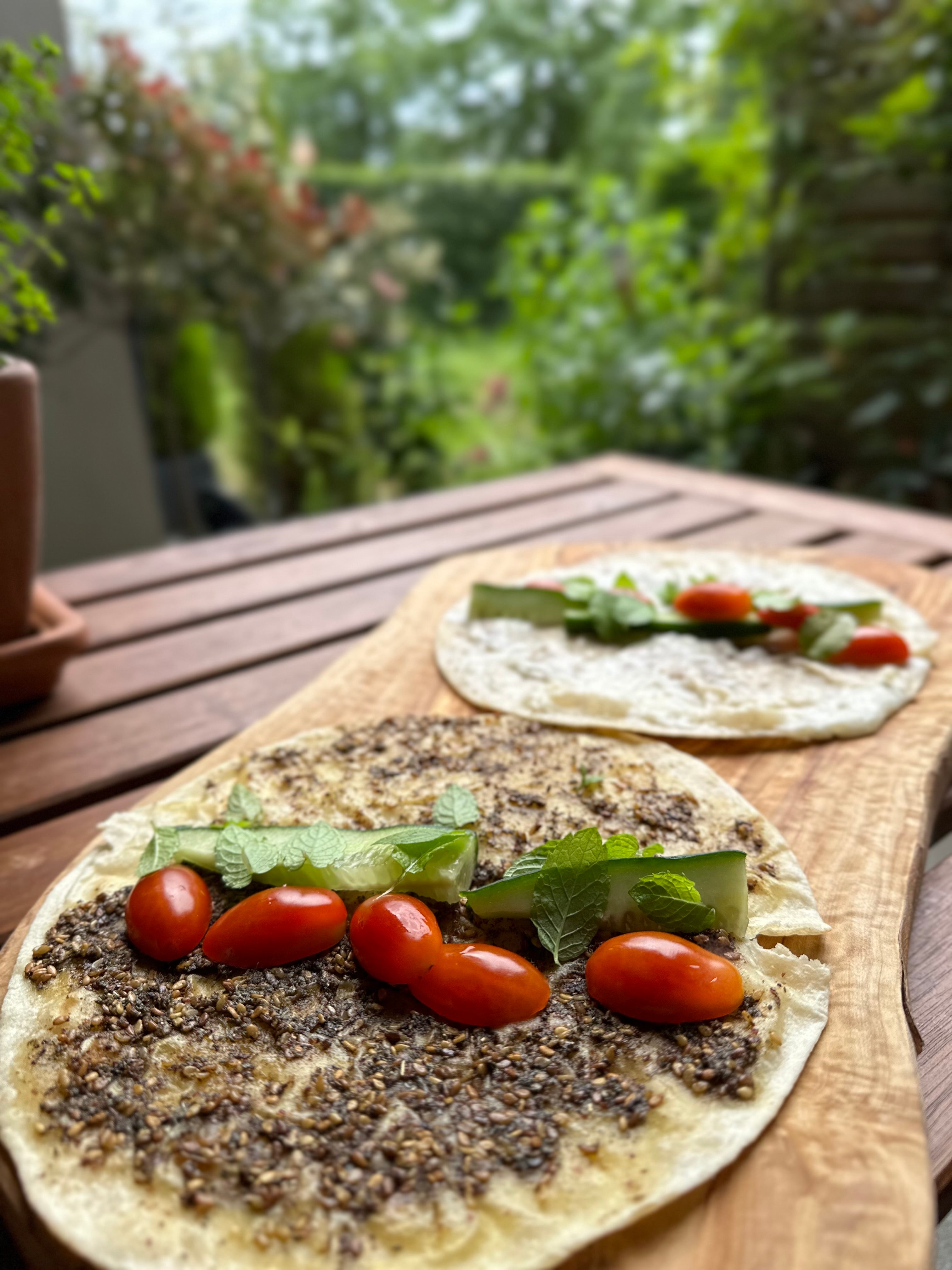 Frisches libanesisches Manakish mit Za'atar, garniert mit Gurke, Minze und Kirschtomaten, serviert auf einem Holzteller im Freien