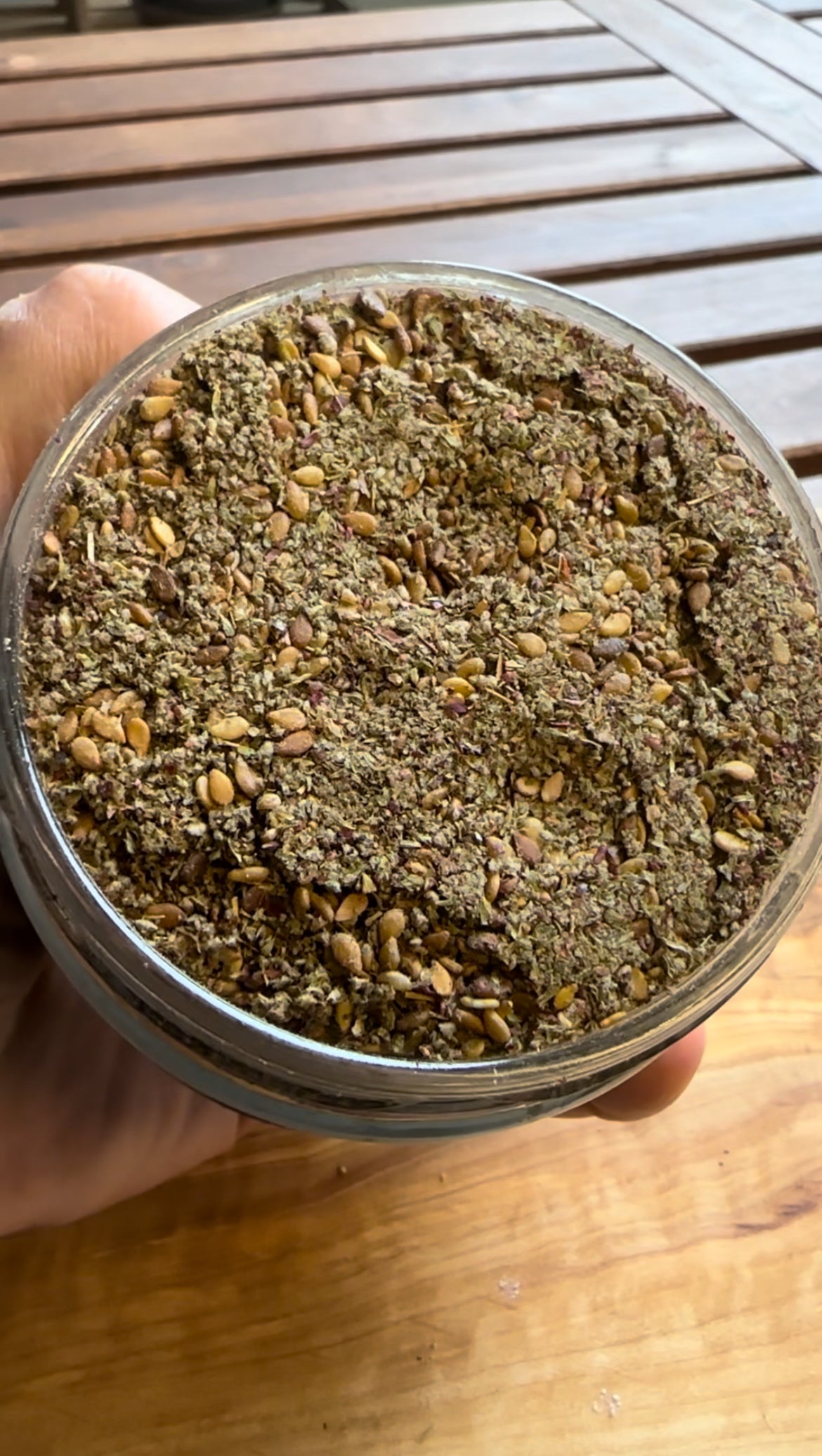 Nahaufnahme eines Glases mit Za'atar-Gewürzmischung, bestehend aus getrockneten Kräutern, Sesamsamen und Gewürzen, gehalten über einem Holztisch