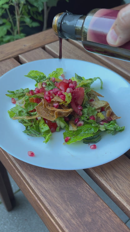 Ein farbenfroher Teller mit Fattoush-Salat, einem klassischen orientalischen Gericht, wird mit einem feinen Strom "honeydew Granatapfelmelasse" veredelt. Die Zutaten – frischer Römersalat, saftige Tomaten, knuspriges Brot, Gurken und leuchtende Granatapfelkerne – präsentieren sich auf einem weißen Teller, der auf einem Holztisch im Freien steht. Die tiefdunkle Melasse fließt elegant über den Salat und verspricht ein harmonisches Zusammenspiel von süß und herzhaft. Der natürliche Hintergrund mit grünem Laub 